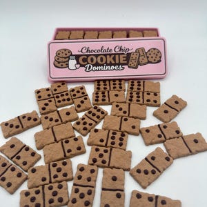 May include: A set of chocolate chip cookie dominoes with a pink box. The dominoes are brown with dark brown chocolate chip dots. The box has the words "Chocolate Chip Cookie Dominoes" on it.