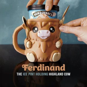 May include: A brown ceramic Highland cow-shaped mug, designed to hold a pint of ice cream. The mug has a handle, two spoons, and the text "Ferdinand THE ICE PINT HOLDING HIGHLAND COW". The ice cream container is blue with the Ben & Jerry's logo.