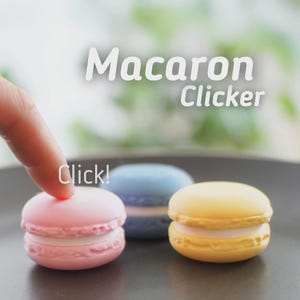 May include: Three colorful macaron-shaped clickers in pink, blue, and yellow, with white filling, are displayed on a dark plate. The word "Macaron Clicker" is written in white, and a finger is about to press the pink one. The word "Click!" is also visible.