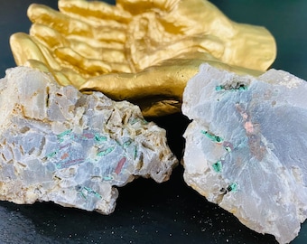 May include: Two pieces of rough, white, and gray agate with green and copper inclusions. The agate is sitting on a gold-painted hand sculpture.