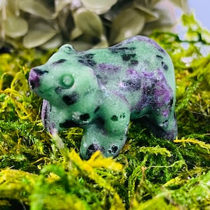 May include: A green and purple carved jade bear figurine with black spots. The bear is standing on a bed of green moss.
