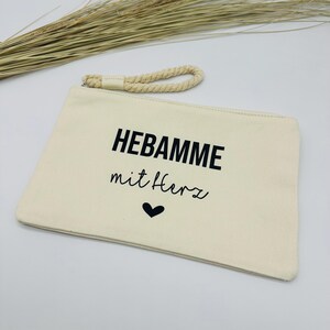 Könnte beinhalten: Cremefarbene Canvas-Tasche mit Kordelgriff. Auf der Tasche steht in schwarzer Schrift "HEBAMME mit Herz", darunter ein schwarzes Herz. Die rechteckige Tasche hat einen Reißverschluss.