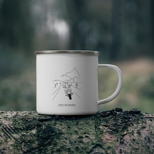 Peut inclure: Mug de camping en émail blanc avec un dessin au trait noir d'une personne conduisant une moto sur une route avec des montagnes en arrière-plan. Le texte "Coffee for Explorers" est imprimé sur le mug.