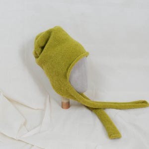 May include: A chartreuse green felted hat with a pointed crown and two long, flat ties extending from the sides. The hat features a soft, textured surface and a rounded opening designed to frame the face, showcasing a simple, functional construction.