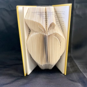 May include: A book folded into the shape of a butterfly. The book is open and the pages are folded to create the butterfly's wings. The book is yellow with white pages.