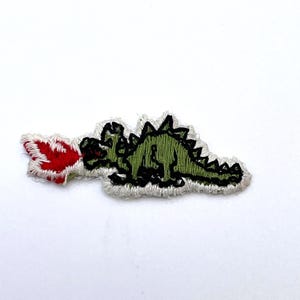 May include: An embroidered patch featuring a green dinosaur with black outlines, depicted in a side profile. The dinosaur has a spiky back and is shown exhaling a bright red and white flame from its mouth, all set against a white background.