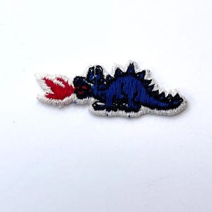 Peut inclure: Un écusson brodé thermocollant représentant un dinosaure bleu foncé au dos épineux, orienté vers la gauche et crachant une flamme rouge et blanche vive. Le motif est entièrement bordé d'un contour blanc épais pour un contraste marqué.