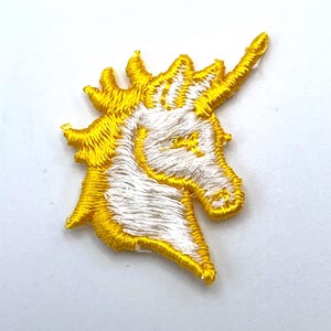 May include: An embroidered patch featuring the head of a unicorn in profile. The design is rendered in bright yellow and white thread, showcasing a prominent horn, a flowing mane, and a detailed eye, all outlined with a thick yellow border.