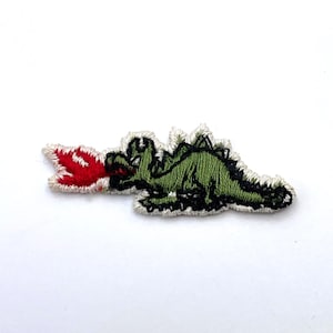 Peut inclure: Un écusson brodé représentant un dragon vert aux contours noirs, crachant une flamme rouge vif et blanche. Le dragon est représenté de profil, avec une bordure brodée blanche entourant toute la forme de la créature et de la flamme.