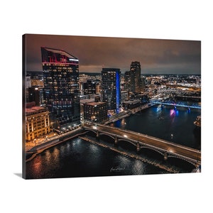 May include: An aerial view of a city skyline at night. The image shows a river with a bridge crossing it. The buildings are lit up with warm and cool lights. The sky is a dark blue with a few clouds.