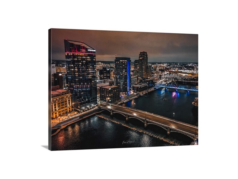 May include: An aerial view of a city skyline at night. The image shows a river with a bridge crossing it. The buildings are lit up with warm and cool lights. The sky is a dark blue with a few clouds.