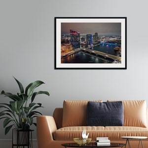May include: An aerial nighttime view of a city skyline with a river and bridge. The buildings are lit up in various colors, including red, blue, and white. The river is reflecting the city lights.