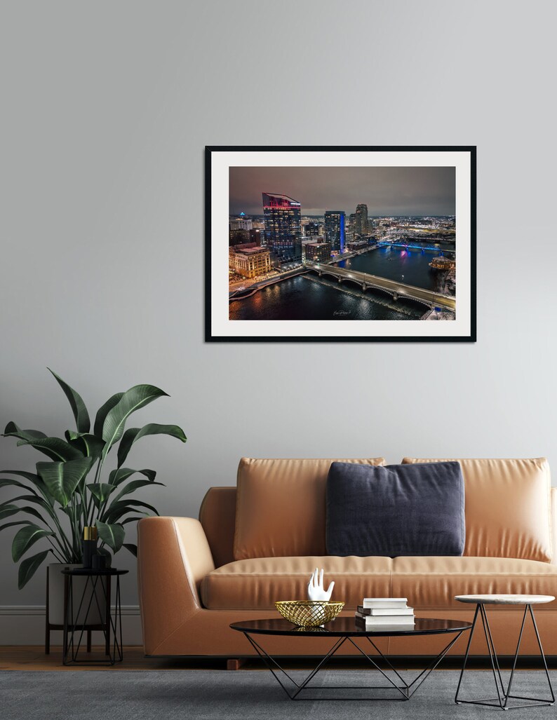 May include: An aerial nighttime view of a city skyline with a river and bridge. The buildings are lit up in various colors, including red, blue, and white. The river is reflecting the city lights.