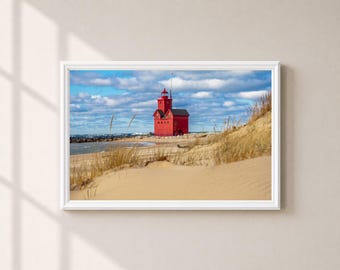 May include: A framed landscape photograph featuring a bright red lighthouse situated on a sandy beach with dunes covered in dry grass. The scene includes a blue sky with scattered white clouds above the water and a rocky shoreline in the distance.
