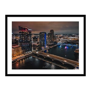 May include: An aerial view of a city skyline at night. The image shows a river with a bridge crossing it. The city is lit up with lights, and there are several tall buildings in the background.