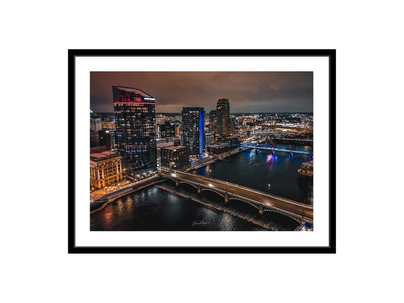 May include: An aerial view of a city skyline at night. The image shows a river with a bridge crossing it. The city is lit up with lights, and there are several tall buildings in the background.