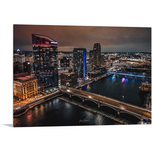 May include: An aerial view of a city skyline at night. The city is lit up with lights, and there is a river running through the center of the image. A bridge crosses the river, and there are buildings on both sides of the river.