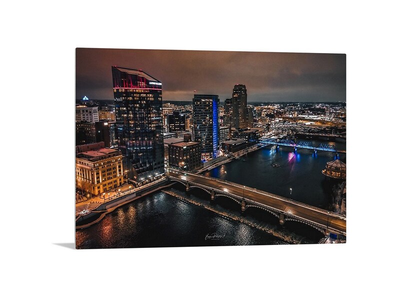 May include: An aerial view of a city skyline at night. The city is lit up with lights, and there is a river running through the center of the image. A bridge crosses the river, and there are buildings on both sides of the river.