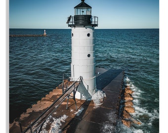 May include: A white lighthouse with a black roof and lantern stands on a rocky pier. Waves crash against the pier, creating a dramatic scene. The sky is a clear blue.