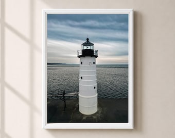 May include: A white cylindrical lighthouse with a black lantern room and small circular windows stands on a dark concrete pier. The structure is positioned against a vast body of water under a cloudy, overcast sky, captured in a vertical frame.