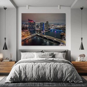 May include: A nighttime aerial view of a city skyline with a bridge crossing a river. The city is lit up with lights, and the river reflects the lights. The image is framed by a bedroom with a bed, nightstands, and lamps.