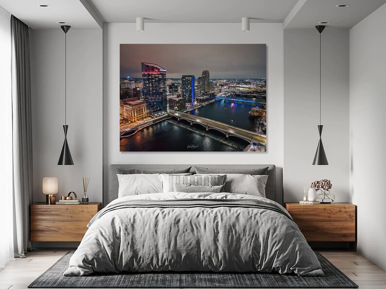 May include: A nighttime aerial view of a city skyline with a bridge crossing a river. The city is lit up with lights, and the river reflects the lights. The image is framed by a bedroom with a bed, nightstands, and lamps.