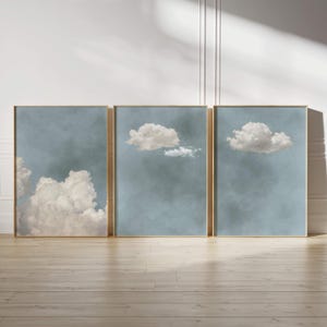 Könnte beinhalten: Drei gerahmte Kunstdrucke mit Wolkenformationen vor einem gedämpften blauen Hintergrund. Die Wolken sind in Weiß- und Beigetönen dargestellt und haben ein weiches, strukturiertes Aussehen. Die Rahmen haben eine goldene Oberfläche, die dem Kunstwerk einen Hauch von Eleganz verleiht.