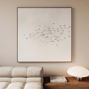 Puede incluir: Obra de arte enmarcada cuadrada que representa una bandada de pájaros negros en vuelo contra un fondo pálido. La obra de arte se exhibe sobre un sofá beige y una mesa auxiliar de madera con una lámpara blanca y un libro.