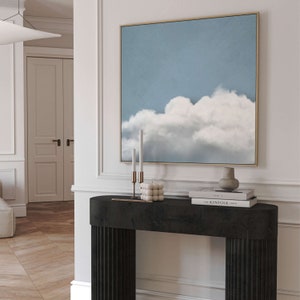 Op de afbeelding: Een zwarte consoletafel met gegroefde poten, met een ingelijst schilderij van een witte wolk tegen een blauwe lucht, twee kandelaars met witte kaarsen, een decoratieve keramische vaas en twee boeken met de titels "Issue 8" en "The Kinfolk Table".