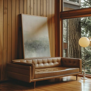May include: A brown leather sofa with a light brown wood frame sits in a room with wood paneling and large windows. A framed abstract painting hangs on the wall behind the sofa. A floor lamp with a white globe shade stands in the corner of the room.
