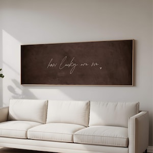 May include: A horizontal rectangular wall art piece featuring a dark brown, textured background with the white cursive text "how lucky are we" followed by a small heart symbol. The artwork is enclosed in a thin, light-colored wooden frame.