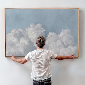 Puede incluir: Una impresión artística enmarcada que presenta un cielo nublado con un fondo azul claro. La obra de arte está en un marco de madera natural. La persona sostiene la impresión enmarcada contra una pared blanca.