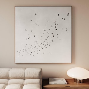 May include: A framed canvas print depicting a flock of birds in silhouette against a pale background. The artwork has a minimalist style, with an emphasis on the birds' movement and the contrast between the dark shapes and the light backdrop. The frame is a neutral colour.