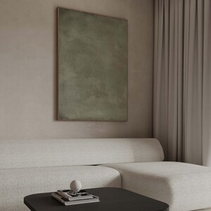 May include: An abstract olive green painting in a wooden frame hangs above a white sofa. A black coffee table holds a stack of books and a white sphere. The room has a neutral color palette with a modern aesthetic.