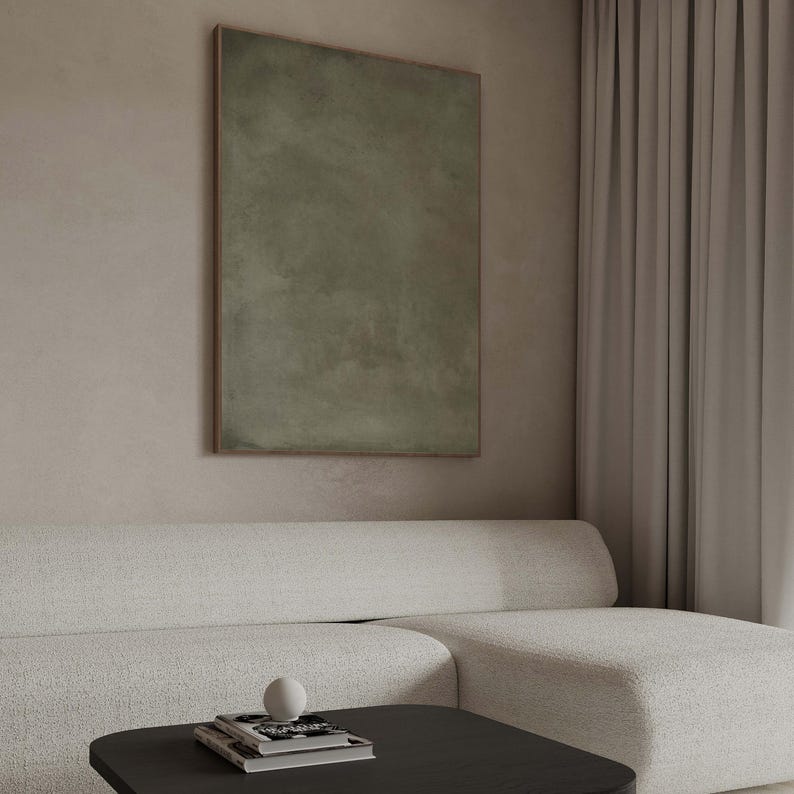 May include: An abstract olive green painting in a wooden frame hangs above a white sofa. A black coffee table holds a stack of books and a white sphere. The room has a neutral color palette with a modern aesthetic.