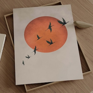 Op de afbeelding: Een ingelijste kunstprint met een minimalistisch ontwerp. Een grote oranje zon op de achtergrond, met een zwerm donkerblauwe vogels die door de lucht vliegen. Het kunstwerk wordt weergegeven in een lichtgekleurde houten lijst, wat een serene en natuurlijke uitstraling cre&euml;ert.