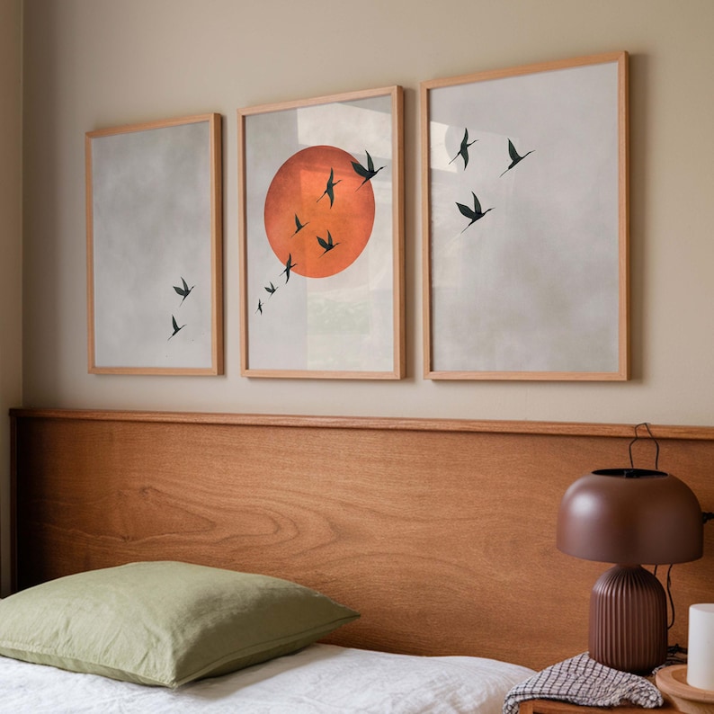 Op de afbeelding: Drie ingelijste kunstdrukken met een minimalistisch ontwerp. Elke print heeft een lichte achtergrond met een vlucht zwarte vogels in vlucht. De centrale print bevat een grote oranje zon. De frames zijn gemaakt van lichtgekleurd hout.