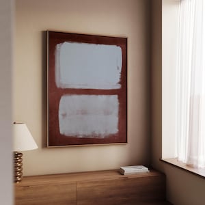 Op de afbeelding: Abstracte kunstprint met twee lichtblauwe rechthoeken op een donkerrode achtergrond, goud omlijst. Het kunstwerk hangt aan een beige muur boven een houten kast, wat een minimalistische esthetiek creëert.