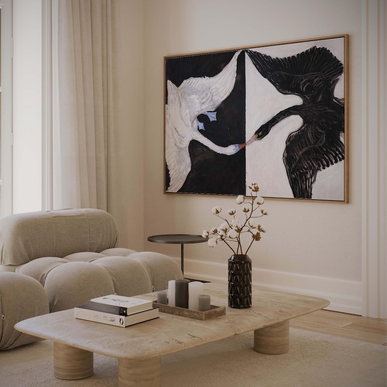 May include: A living room interior with a modern couch, a coffee table, and a black and white painting of two swans with their beaks touching. The painting is framed and hanging on the wall. There is a vase with cotton flowers on the table.