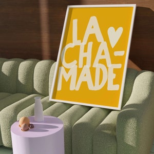 May include: A framed print with a yellow background and white text that reads "LA CHA-MADE" with a white heart symbol. The print is hanging on a wall above a green sofa.