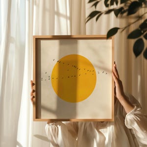 以下が含まれることがあります： 黄色い太陽と、その周りを飛ぶ黒い鳥の群れが描かれた額装されたプリント。プリントは白い背景に印刷され、明るい木製のフレームで額装されています。