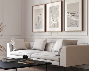 May include: A white sofa with three pillows sits in front of a white wall with three framed abstract art prints. The prints are in shades of brown and black with thin lines and shapes. The sofa is in a living room setting with a black coffee table in front of it.