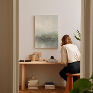 May include: A woman sits at a desk with a light wood top and a stack of books. A painting with a green and blue abstract design hangs on the wall behind her. There are several small ceramic pots and a wooden tray on the desk.