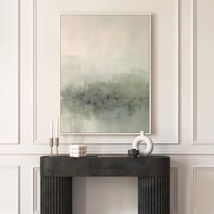 May include: A framed abstract painting with a muted green and gray color scheme hangs above a black console table. The painting depicts a hazy landscape with a soft, ethereal feel. The console table has a textured surface and features a white ceramic vase and a black decorative object.