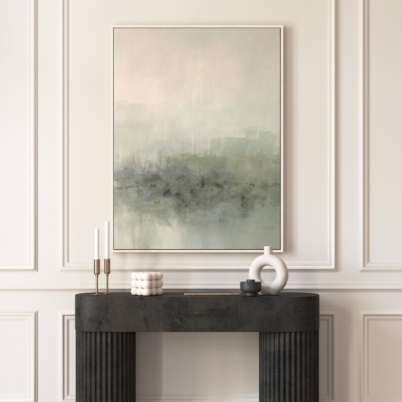 May include: A framed abstract painting with a muted green and gray color scheme hangs above a black console table. The painting depicts a hazy landscape with a soft, ethereal feel. The console table has a textured surface and features a white ceramic vase and a black decorative object.