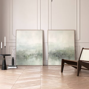 May include: Two abstract landscape paintings in muted green and white tones, hanging on a white wall in a modern interior. The paintings are framed in a light wood frame. A wooden chair with a woven seat is in the foreground.