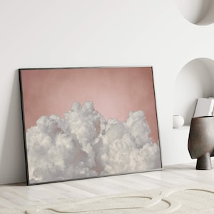 May include: Framed art print featuring a cloudscape. The artwork showcases fluffy white clouds against a soft pink background. The print is encased in a black frame, adding a modern touch to the serene scene. The artwork is leaning against a white wall.