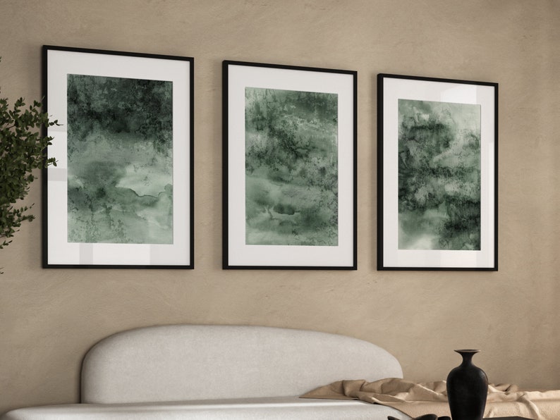 K&ouml;nnte beinhalten: Drei gerahmte abstrakte Kunstdrucke. Jeder Druck zeigt ein Design im Aquarellstil in Gr&uuml;n- und Wei&szlig;t&ouml;nen, das in einem schwarzen Rahmen eingefasst ist. Das Kunstwerk wird &uuml;ber einem wei&szlig;en Sofa ausgestellt und schafft eine moderne &Auml;sthetik.