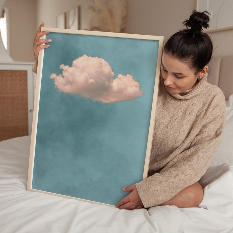 May include: A framed print of a single, fluffy, pink cloud against a blue sky. The print is being held by a person in a beige sweater.