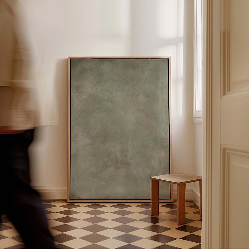 May include: A large, rectangular framed artwork with a muted green textured surface. A small, square wooden stool sits beside the artwork. The floor has a black and white checkered pattern. A person is blurred in the foreground.